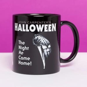 Puede incluir: Una taza de cerámica negra con el texto blanco John Carpenter's HALLOWEEN y The Night He Came Home! junto a una ilustración gráfica de una mano sosteniendo un cuchillo grande. La taza tiene un asa curva y un acabado brillante.