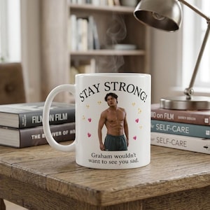 Könnte beinhalten: Ein weißer Keramikbecher mit dem schwarzen Text STAY STRONG! oben und Graham wouldn't want to see you sad. unten. In der Mitte befindet sich ein Fotodruck einer oberkörperfreien Person in dunklen Shorts, umgeben von kleinen rosa Herzen.