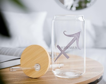 May include: A clear glass can-shaped tumbler with a bamboo lid and straw. The tumbler features a Sagittarius zodiac symbol and text in a soft purple hue. The lid is round and made of light-colored bamboo. The glass is sitting on a wooden table.