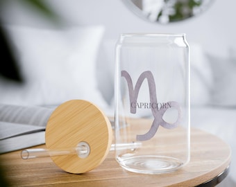 May include: A clear glass tumbler with a bamboo lid and straw. The tumbler features the Capricorn zodiac symbol and the word "CAPRICORN" in a light purple color. The tumbler is sitting on a wooden table.