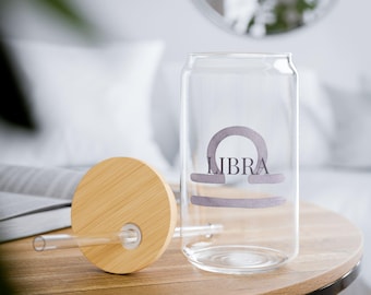 May include: A clear glass can-shaped tumbler with a bamboo lid and straw. The tumbler features the word "LIBRA" and a Libra symbol in a light gray color. The tumbler sits on a wooden table.