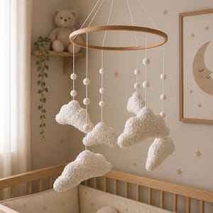 Puede incluir: Un móvil colgante con un marco circular de madera suspendido por cuerdas blancas. Del marco cuelgan varios elementos de felpa blancos y texturizados en forma de nube, intercalados con pequeñas cuentas esféricas blancas a distintas alturas.