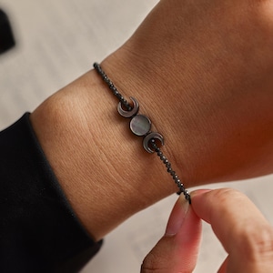 Op de afbeelding: Een verfijnde armband met een centrale bedel bestaande uit een ronde schijf geflankeerd door twee maansikkels. De bedel is gemaakt van een donker, iriserend materiaal. De band bestaat uit kleine, gefacetteerde donkere kralen aan een dun koord.