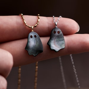Op de afbeelding: Twee spookvormige hangers gemaakt van iriserend donker schelpmateriaal. De ene hanger hangt aan een goudkleurige kralenketting, terwijl de andere aan een zilverkleurige schakelketting hangt. Elke spookhanger heeft twee ronde uitsparingen als ogen.