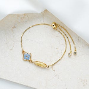 May include: Gold-coloured adjustable bracelet with a light blue flower and a gold oval accent. The bracelet has a delicate chain design with two small, clear, round charms at the end of the adjustable pull strings. The bracelet is on a light-coloured surface.