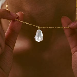 Könnte beinhalten: Eine goldene Halskette mit einem weißen, schillernden, geisterförmigen Anhänger aus Perlmutt. Der Anhänger hat zwei kleine, kreisförmige Aussparungen für die Augen und hängt an einer zarten Gliederkette aus goldfarbenem Metall.