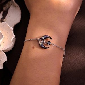 Könnte beinhalten: Ein silbernes Kettenarmband mit einem Halbmond-Anhänger. Der schwarze Mond-Anhänger ist mit getrockneten Blumen gefüllt und hat einen schwarzen Edelstein in der Mitte. Das Armband wird am Handgelenk getragen.