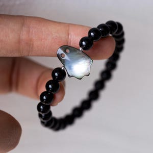 Könnte beinhalten: Ein elastisches Armband mit einer Reihe glatter, runder, polierter schwarzer Perlen. Der Blickfang ist ein flacher, schillernder, grauer Muschelanhänger in Form eines Geistes mit zwei kreisförmigen Aussparungen für die Augen.