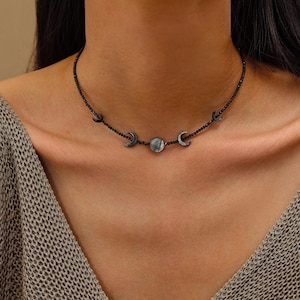 Op de afbeelding: Een zwarte kralen choker ketting met een centraal rond hangertje met een parelmoer afwerking, geflankeerd door twee kleine maansikkelbedels aan weerszijden. De ketting is gemaakt van kleine, donkere, uniforme kralen in een enkele streng.