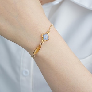 Può includere: Un delicato braccialetto dorato con un fiore azzurro e una pietra oblunga color oro-marrone. Il braccialetto è indossato su un polso, mettendo in risalto la sua catena sottile e il suo design. Il fiore è incastonato in una cornice dorata, aggiungendo un tocco di eleganza.