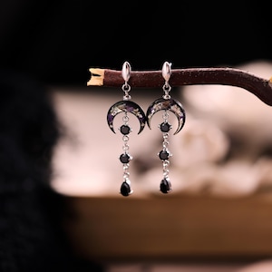 May include: A pair of silver-toned dangle earrings featuring a crescent moon charm filled with small purple and white dried flowers. Below each moon, two round black stones and one teardrop-shaped black stone hang from a silver-toned chain.