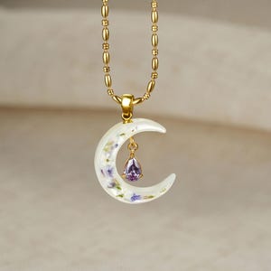Può includere: Una collana a catena con perline dorate caratterizzata da un ciondolo a mezzaluna realizzato in materiale bianco traslucido con piccoli fiori secchi incorporati. Una singola gemma viola a forma di pera pende dal centro della luna.