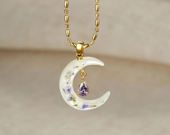 May include: A gold-toned beaded chain necklace featuring a crescent moon pendant made of translucent white material embedded with small dried flowers. A single pear-shaped purple gemstone hangs from the center of the crescent moon charm.