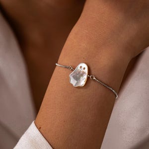 Op de afbeelding: Een delicate zilverkleurige kettingarmband met een centrale bedel gesneden uit iriserend parelmoer in de vorm van een geest met twee ronde uitsparingen voor de ogen. De armband bevat een dunne, verstelbare metalen schakelketting.
