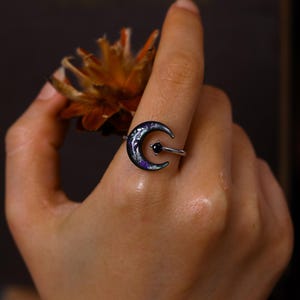 May include: A silver-toned ring featuring a crescent moon shape with a dark, speckled finish containing purple and silver flecks. A small, round, black gemstone is set in the center of the crescent moon, attached to a thin, polished silver band.