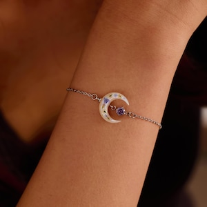 May include: A delicate silver-toned chain bracelet featuring a crescent moon charm made of white resin embedded with tiny dried blue and green flowers, accented by a single small round purple gemstone suspended within the curve of the moon.