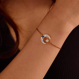 May include: A delicate gold-tone chain bracelet featuring a crescent moon charm. The moon charm is white with embedded blue floral accents and a single clear round gemstone set in the centre of the crescent curve, attached to a thin metal chain.