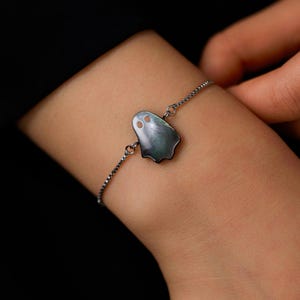 May include: A delicate bracelet featuring a dark, iridescent ghost-shaped charm made of mother-of-pearl. The charm has two small circular cutouts for eyes and is attached to a thin, dark-toned metal chain with a classic box link design.