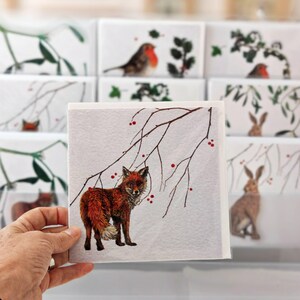 Peut inclure: Une carte carrée avec une illustration détaillée d'un renard roux, debout sous une branche avec des baies rouges. D'autres cartes avec des illustrations similaires sur le thème de la nature sont visibles en arrière-plan.