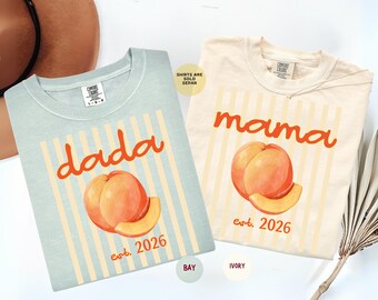 May include: A light blue t-shirt with the text "dada est. 2026" and an ivory t-shirt with the text "mama est. 2026". Both shirts feature a peach graphic and vertical stripes. The shirts have a Comfort Colors label at the inner neckline.