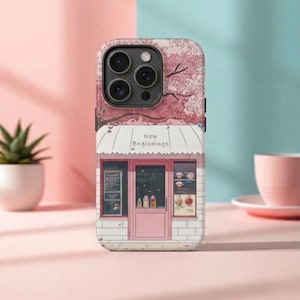 Op de afbeelding: Telefoonhoesje met een roze winkelontwerp en de tekst "New Beginnings" onder een kersenbloesemmotief. De hoes heeft een roze, witte en blauwe kleurstelling, met een kleine plant en een roze kopje op de achtergrond.