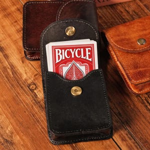 Può includere: Astuccio per carte in pelle nera con chiusura a scatto, contenente un mazzo di carte da gioco Bicycle. L'astuccio è aperto, rivelando il design rosso e bianco delle carte. Altri astucci in pelle sono sullo sfondo, su una superficie di legno.