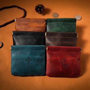 Può includere: Una collezione di portamonete in pelle in vari colori, tra cui marrone, verde, nero e rosso. I portamonete hanno una forma rettangolare con una chiusura in metallo. Uno ha una croce e il nome "Charlotte", un altro la lettera "G".