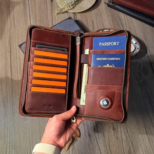 Könnte beinhalten: Geöffnete braune Reisebrieftasche aus Leder mit mehreren Kartenfächern, orangefarbenen Karten und einem Reißverschluss. Die Brieftasche enthält einen Reisepass, Bargeld und ein Apple AirTag.