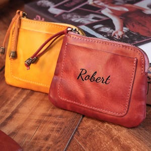 May include: Two leather coin purses, one in a vibrant yellow and the other in a reddish-orange. The reddish-orange purse has the name "Robert" printed on it. Both purses have a zipper closure and a small pocket on the front.
