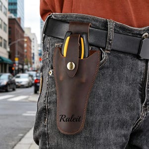 Op de afbeelding: Een donkerbruine lederen holster met een drukknoopsluiting en de tekst Raleit op de voorzijde gedrukt. De holster is bevestigd aan een zwarte riem en bevat een gereedschap met gele en zwarte handvatten, gedragen op een grijze spijkerbroek.
