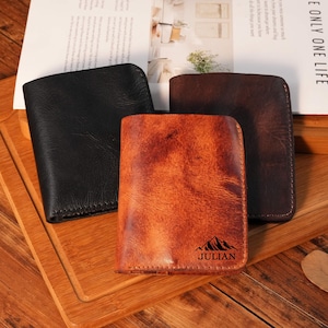 May include: Three leather wallets in black, brown, and tan are displayed on a wooden tray. The tan wallet has a mountain graphic and the name "JULIAN" printed on it. The wallets are rectangular and appear to be bi-fold.