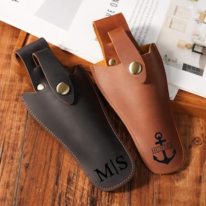 Op de afbeelding: Twee lederen holsters met riemlussen en koperen drukknoopsluitingen. Het donkerbruine holster heeft de letters M|S onderaan gegraveerd. Het lichtbruine holster heeft een ankergrafiek met het woord HUNTER boven het anker gegraveerd.