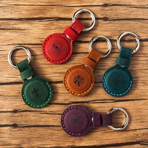 Puede incluir: Un conjunto de llaveros de cuero personalizados en varios colores, incluyendo rojo, verde, naranja, turquesa y morado. Cada llavero presenta un diseño circular con un llavero plateado e iniciales o símbolos en relieve. Sobre una superficie de madera.