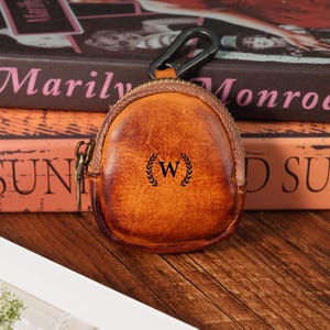 May include: A small, round, brown leather coin purse with a zip closure and a metal carabiner clip. The purse features a black initial "W" inside a laurel wreath. The purse is on a wooden surface.