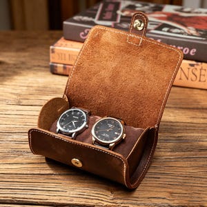 May include: A brown leather watch roll case with a snap button closure, featuring a soft interior lining that holds two wristwatches with black faces and silver-toned metal cases. The exterior leather has visible stitching along the edges.