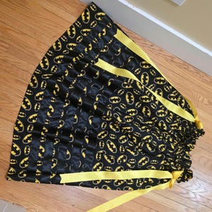 May include: Black satin fabric featuring a repeating yellow Batman logo pattern. The fabric is gathered at the top and tied with yellow ribbons. This item is likely a drawstring bag or decorative piece.