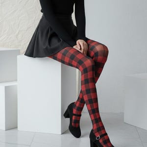 Puede incluir: Zapatos de tacón Mary Jane negros con tacón de bloque y correa en el tobillo. El atuendo incluye una falda negra y una camiseta negra de manga larga. Las medias tienen un estampado de cuadros rojos y negros. El conjunto está sobre un fondo blanco.