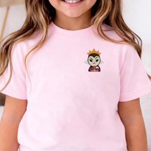 May include: A light pink t-shirt featuring an embroidered cartoon image of a queen with a gold crown. The queen has a white collar and a purple and black dress. The shirt is made of a soft material.