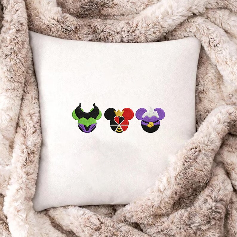 Peut inclure: Coussin carr&eacute; blanc avec des motifs brod&eacute;s de trois visages de personnages de dessins anim&eacute;s. Les motifs pr&eacute;sentent des couleurs noires, vertes, violettes, rouges et jaunes. Le coussin est pos&eacute; sur une couverture en fausse fourrure.