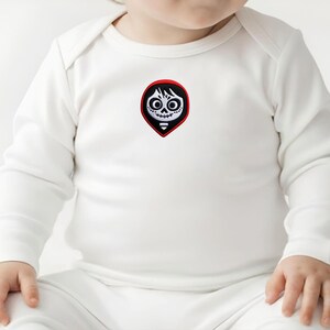 May include: White baby onesie with long sleeves. The onesie features a colorful patch of a stylized skull with black hair, large eyes, and a stitched mouth. The patch is set within a red border.