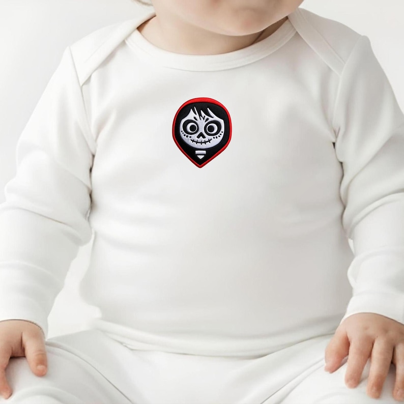 May include: White baby onesie with long sleeves. The onesie features a colorful patch of a stylized skull with black hair, large eyes, and a stitched mouth. The patch is set within a red border.