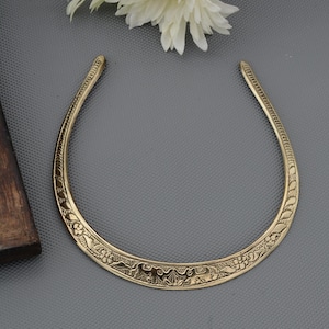 May include: A gold-colored collar necklace with intricate floral and animal carvings. The necklace has a U-shape and is designed to sit around the neck. The details include a series of repeating patterns and a smooth, polished finish. The background is a solid gray.