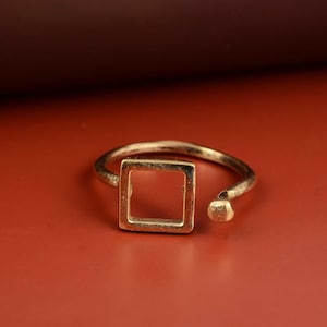 Puede incluir: Un anillo dorado con un centro cuadrado y un diseño abierto. El anillo tiene una forma geométrica simple y un pequeño detalle redondeado en el extremo abierto. El anillo está sobre un fondo rojo.