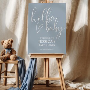 May include: A baby shower sign on a wooden easel. The sign is light blue with the words "hello baby" in cursive, and the text "Welcome to Jessica's Baby Shower" below. A teddy bear and blue fabric are on a wooden stool.