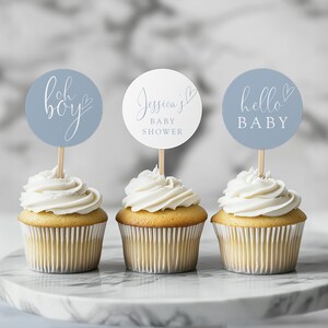Pode incluir: Três cupcakes com cobertura branca e toppers decorativos. Os toppers são redondos, com os textos "Oh boy", "Jessica's BABY SHOWER" e "hello BABY". Os cupcakes estão em um suporte de bolo de mármore.