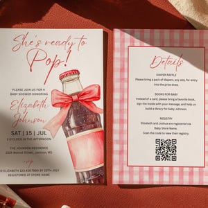 May include: Two rectangular baby shower invitation cards with white backgrounds. The left card features a watercolor soda bottle with a red bow and the text She's ready to Pop! The right card has a pink gingham border and the text Details with a QR code.