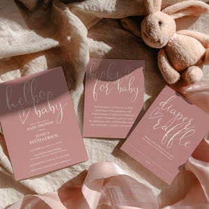 Puede incluir: Tres tarjetas rosas para baby shower con texto de caligrafía blanca, incluyendo "hello baby", "books for baby" y "diaper raffle". Un peluche de conejito descansa cerca sobre un fondo de tela texturizada. Las tarjetas son de color rosa empolvado.