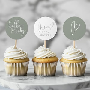 Pode incluir: Três cupcakes com cobertura branca e toppers decorativos. Um topper diz "hello baby" com um coração, outro diz "Jessica's BABY SHOWER", e o último tem um contorno de coração. Os cupcakes estão em um suporte de bolo de mármore.