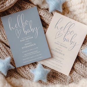 Op de afbeelding: Twee uitnodigingen voor een babyshower, één lichtblauw en één crème, met de woorden "hello baby" in cursief schrift. De uitnodigingen bevatten evenementdetails. Lichtblauwe stervormige decoraties zijn verspreid over een gebreide deken.