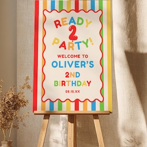 Puede incluir: Un colorido letrero de fiesta de cumpleaños sobre un caballete de madera. El letrero dice "READY 2 PARTY!" y da la bienvenida a los invitados a "OLIVER'S 2ND BIRTHDAY" con la fecha 08.15.XX. El letrero tiene un fondo blanco, un borde rojo ondulado y rayas verticales.